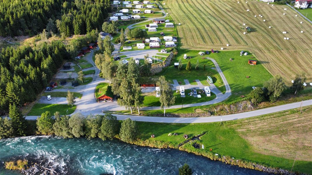 Luftfoto av ein campingplass med bubilar og telt ved ei elv, veg, skogkant og slåtteåkrar med rundballar..