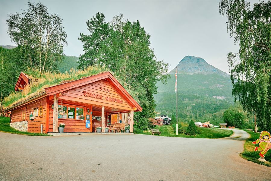 Raudt trebygg med torvtak ved veg, omgitt av grøne tre, med fjell og flaggstong i bakgrunnen..