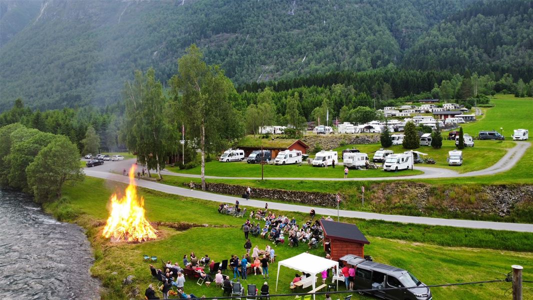 Stort bål ved ei elv med folk samla på graset, campingvogner i bakgrunnen og skogkledde fjell rundt..