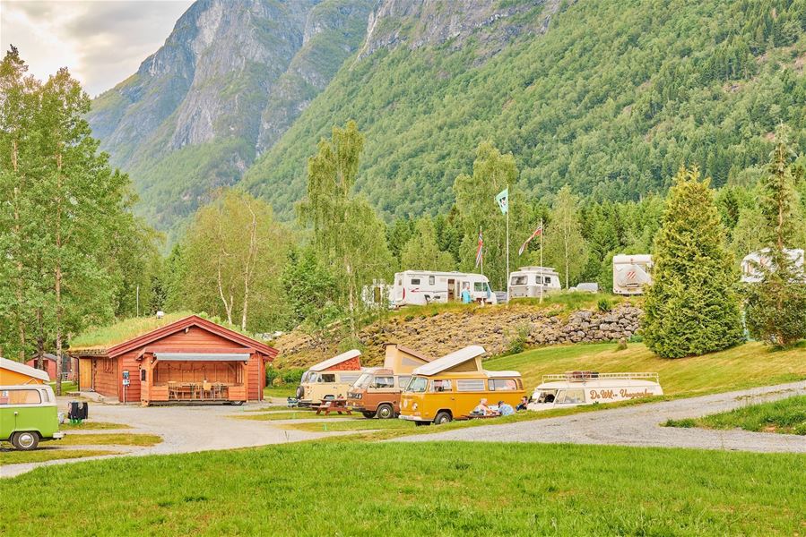 Campingplass med raud hytte, veteranbussar og bubilar i grønt landskap, med skogkledde fjell i bakgrunnen..