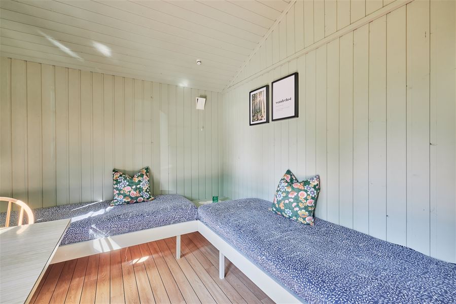 Lite rom med lyse panelveggar, to blå dagbenker i vinkel, blomstrete puter og to innramma bilete på veggen..