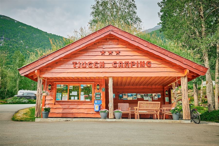 Raudt trebygg merka «Tjugum Camping» med benkar og plakatar under tak, omgitt av tre og fjell..