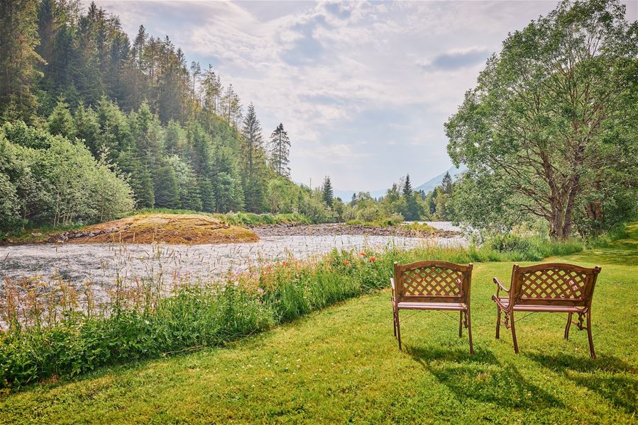 To trebenkar står på ei grasplen ved ei stri elv, med skogkledd åsside og lettskyet himmel i bakgrunnen..