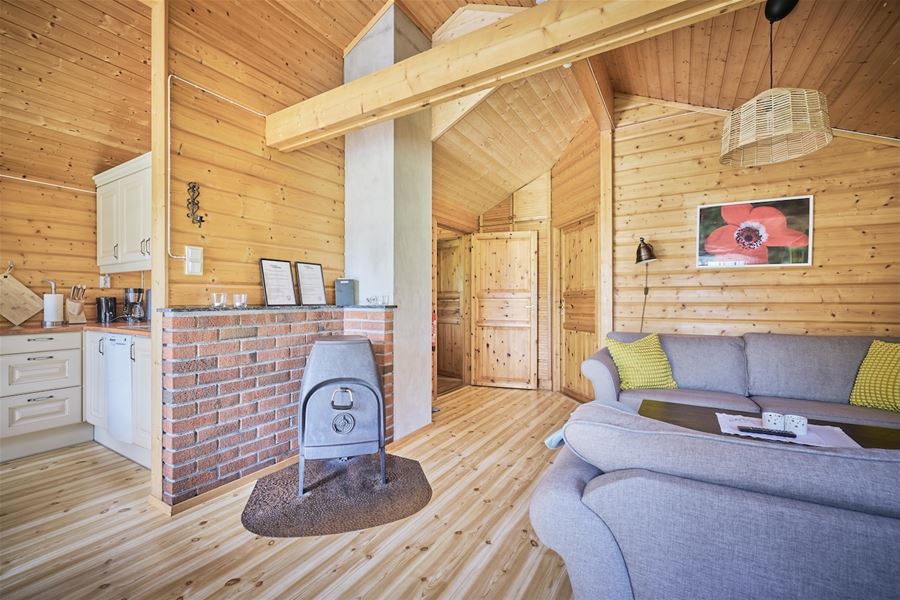 Lys hytte med trepanel, open kjøkenkrok, vedomn ved mursteinsvegg og grå sofa med puter og bilete..