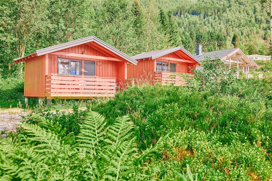 To raude trehytter med små terrassar står i frodig grøne omgjevnader med skogkledd ås i bakgrunnen..