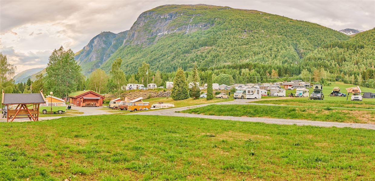 Campingplass med bubil og telt på graseng, med hytter og tett skog framfor eit stort, grønt fjell under overskya himmel..