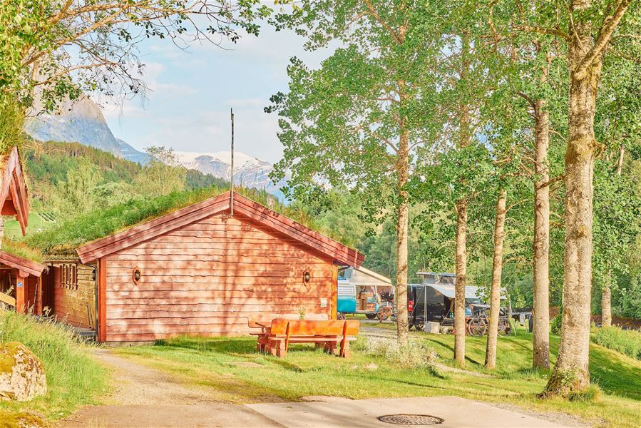 Raud trehytte på grasplen med benk, bjørketrær og campingvogner i bakgrunnen, fjell og blå himmel..