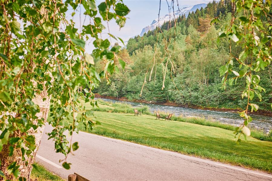 Grøn park ved ei rennande elv med fleire benkar, omkransa av skog og fjell i bakgrunnen..