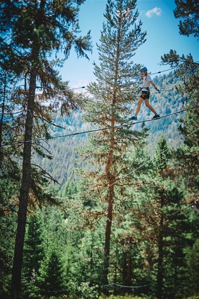 Ein person går på ein stram line mellom høge furutre i fjellskog, med blå himmel og fjerne åsar i bakgrunnen..