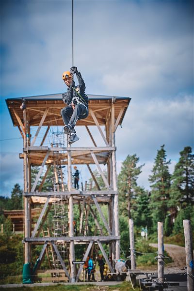 Ein person med hjelm og sele glir i zipline framfor eit høgt tretårn, med skog og blå himmel i bakgrunnen..
