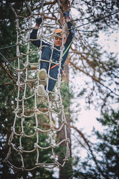 Ein person klatrar i eit tau-nett i ein skogkledd klatrepark, sikra med sele og line mellom trea..