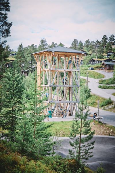 Eit høgt klatretårn av tre med folk i sele, omgitt av furuskog og hytter ved ein svingete veg..