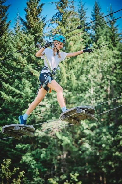 Ein deltakar i klatrepark balanserer på hengande plattformer med hjelm og sele, omgitt av grøne tre..