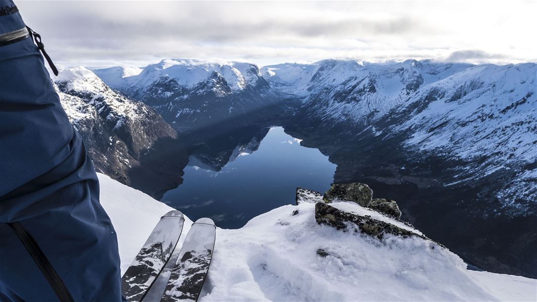 Skier’s legs and ski tips on a snowy ridge above a calm fjord, with rugged snow-covered mountains under clouds..