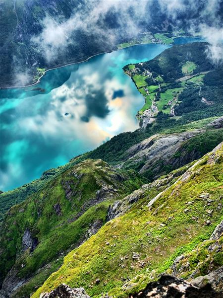 High view from a grassy rocky mountainside over a turquoise lake, with clouds, forested slopes, and small farms..