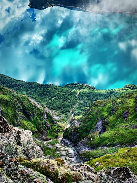 Steep green mountain slopes and rocky ravine descend toward a turquoise lake partly covered by low clouds..