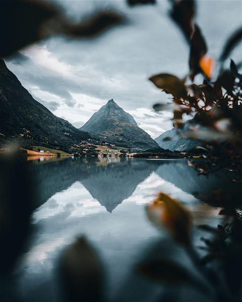 A pointed mountain rises beyond a calm lake, reflecting in the water under cloudy skies, framed by blurred leaves..