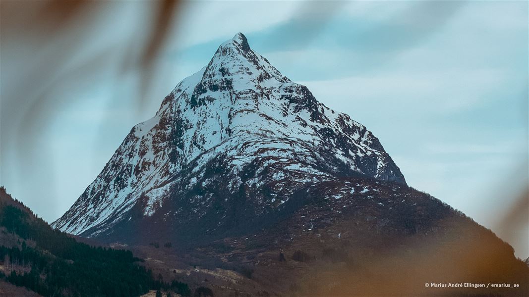 A rugged snow-covered mountain peak rises above dark foothills under a pale blue, lightly clouded sky..