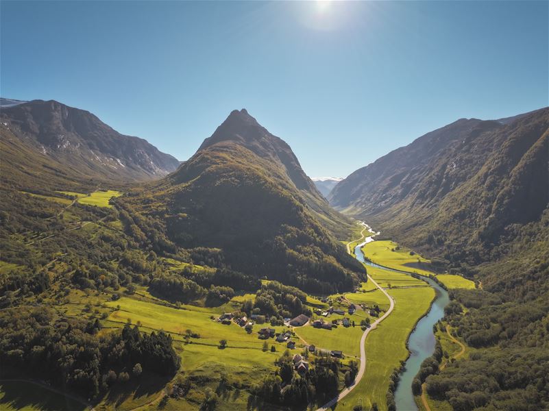 Aerial view of a green valley with a winding river, small village, and steep mountains under a clear blue sky..