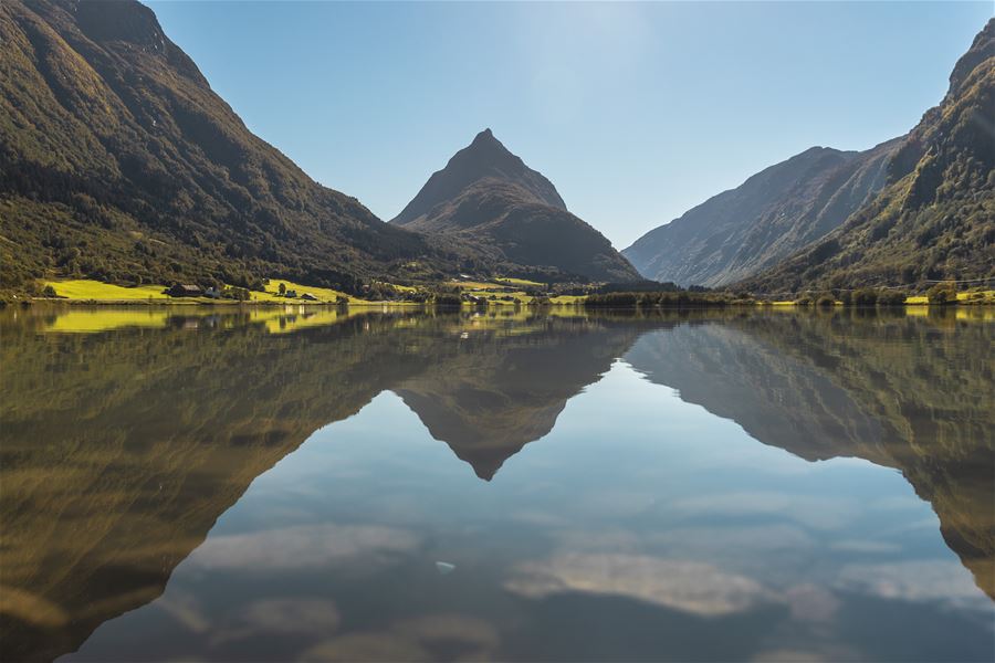 A sharp mountain peak rises between forested slopes, reflected in a still lake under a clear blue sky..