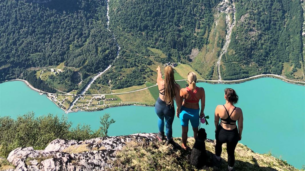 Tre kvinner og ein hund står på ein fjellkant og ser ned på ei turkis innsjø og grøne fjellsider..