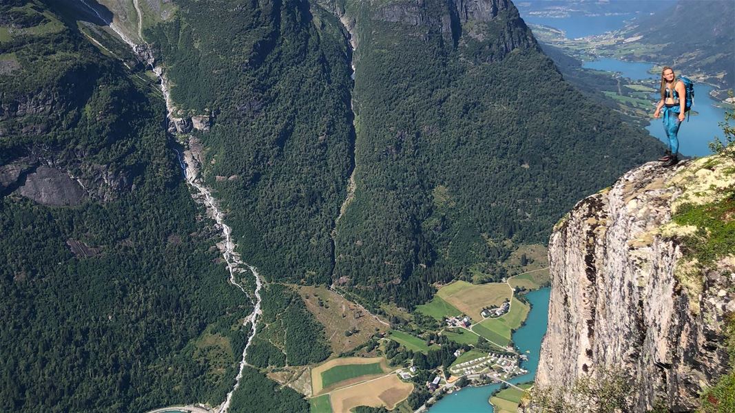 Ein turgåar med ryggsekk står på ein bratt klippekant med utsyn over grøne fjell, dal og blå fjord..