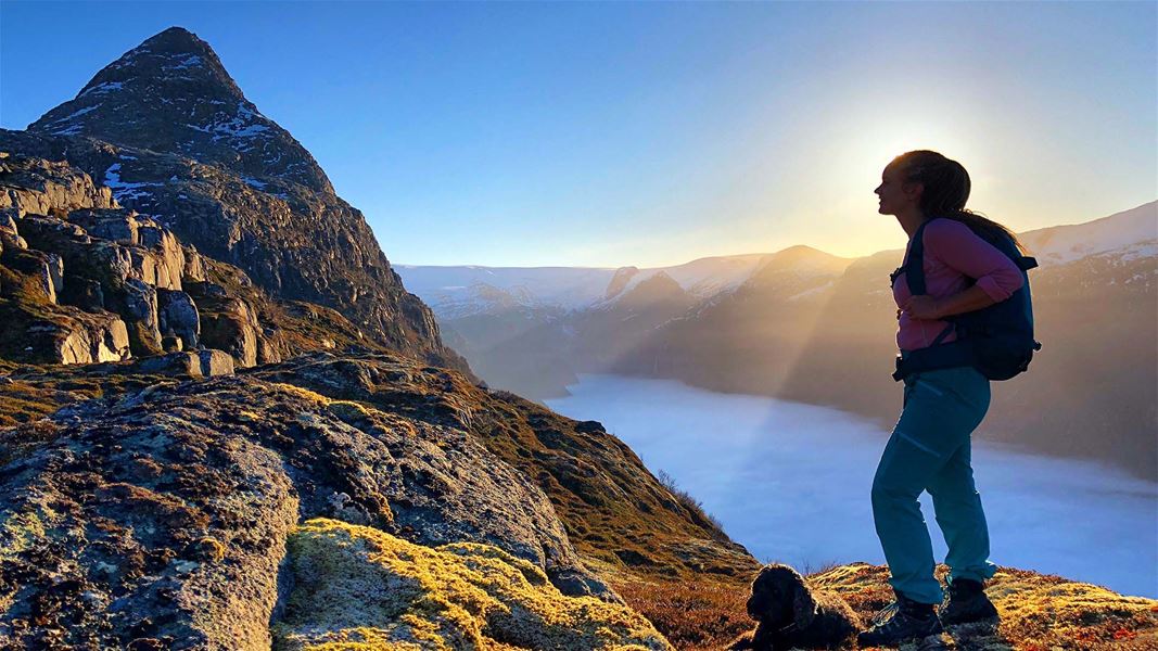 Ein turgåar med ryggsekk står på ein steinete fjellrygg i motlys, med fjord og fjell i bakgrunnen..