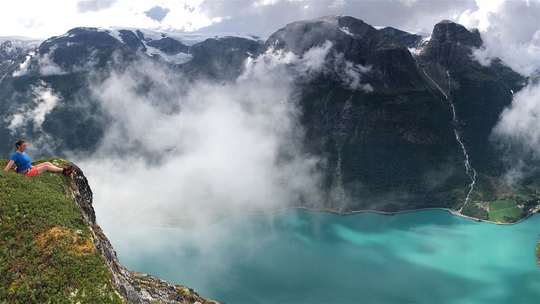 Ein turgåar sit på ein grasdekt klippekant med utsikt mot turkis fjord, skodde og bratte fjell..