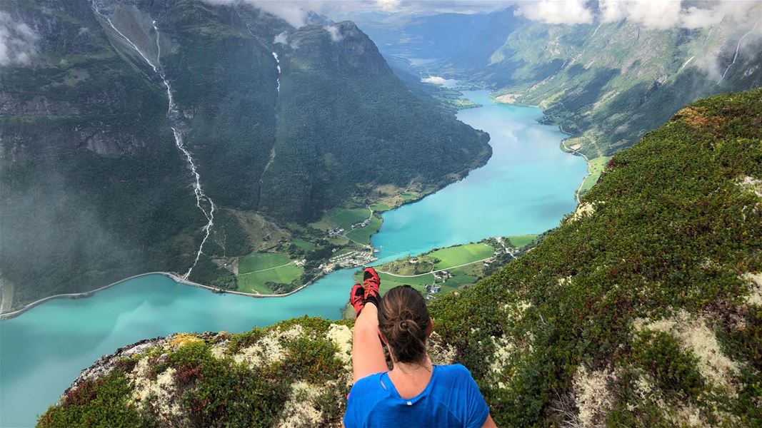 Person sit på fjellkanten og ser ned på ein turkis fjord med grøne dalar, bratte fjell og skyer..