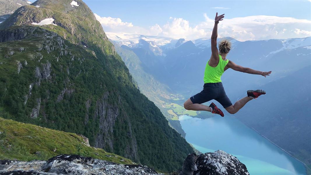 Person i neon grøn topp hoppar frå ein fjellkant med armane ut, med fjord og fjell i bakgrunnen..