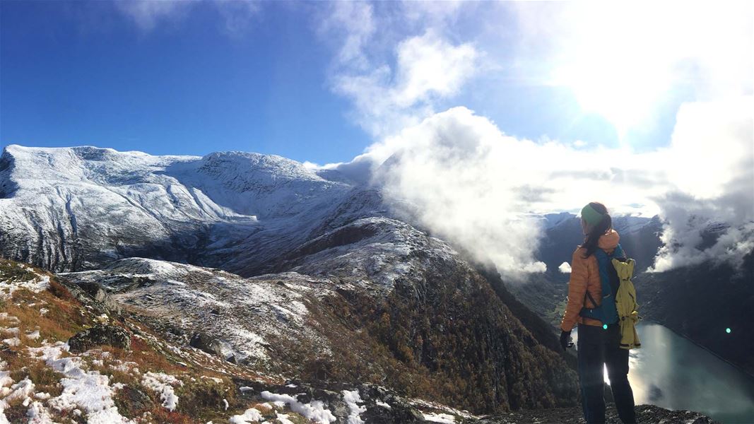 Ein turgåar med ryggsekk står på ein snødekt fjellrygg og ser mot fjell, skyer og fjord i dalen..