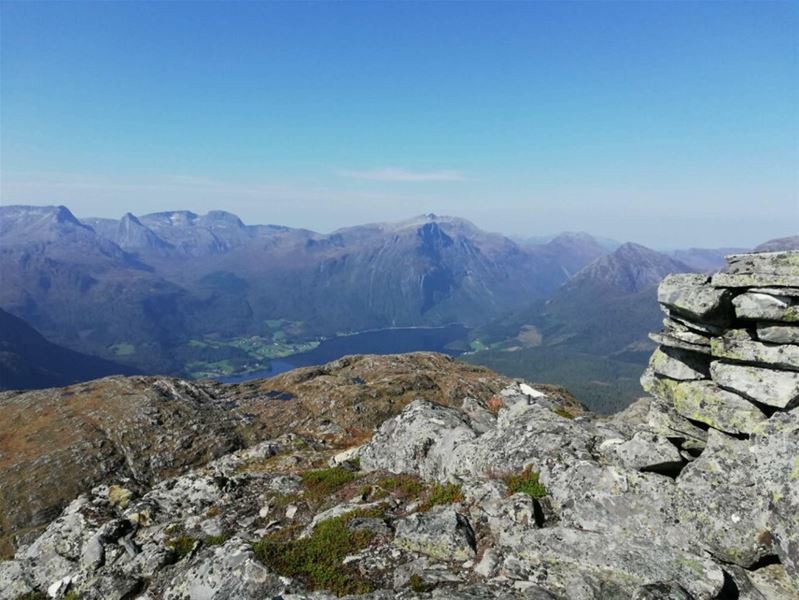 Panoramautsikt frå Helsethornet med steinete forgrunn, fjellrekke og innsjø i dalen under klar blå himmel..
