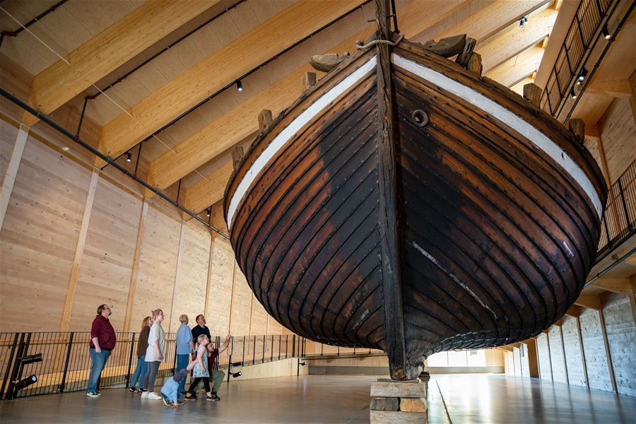Large wooden boat hull displayed indoors on supports, with visitors standing and walking beneath high timber beams..