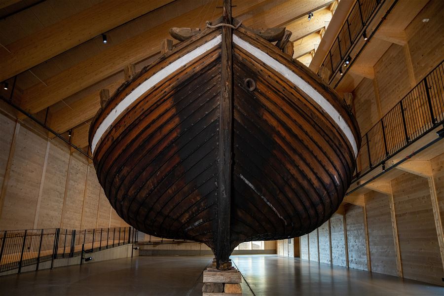 Large wooden boat hull suspended inside a spacious timber-framed hall with railings, spotlights, and polished floor..