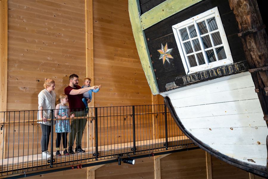 A group of visitors stands on an indoor walkway beside a large wooden boat hull, Holvikejekta, with a window and painted star..
