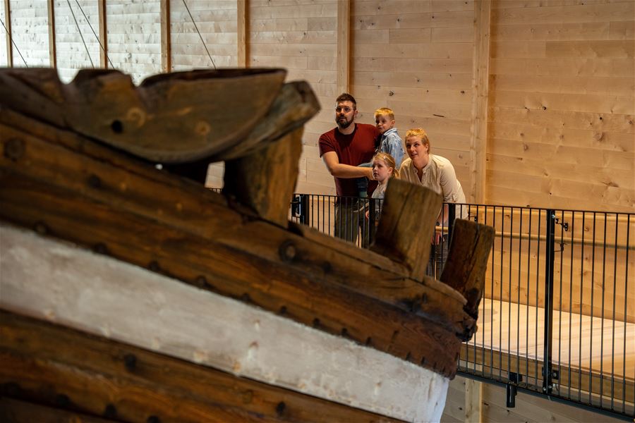 A small group stands behind a railing in a wooden interior, looking toward a large rustic wooden object in front..
