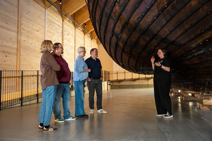 A small group listens to a guide inside a wooden hall beneath a large suspended wooden ship hull..
