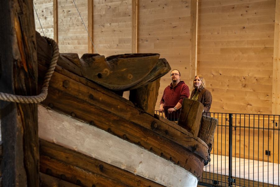 Large wooden ship bow on display indoors, with two visitors standing behind a railing against wood-paneled walls..