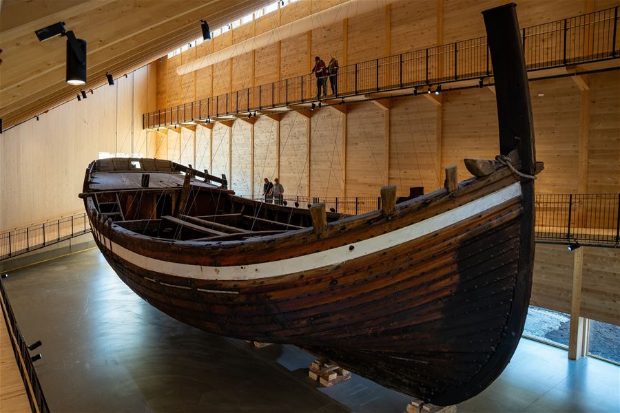 Large wooden boat, Holvikejekta, displayed indoors in a timber museum hall, with elevated walkways and a few visitors above..