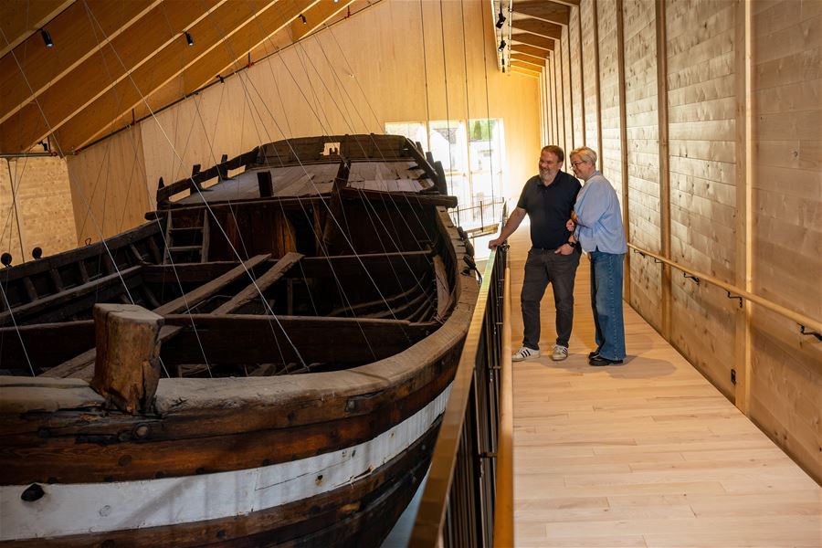 Two visitors stand on a wooden walkway beside a large old wooden boat, Holvikejekta, displayed indoors under a sloped roof..