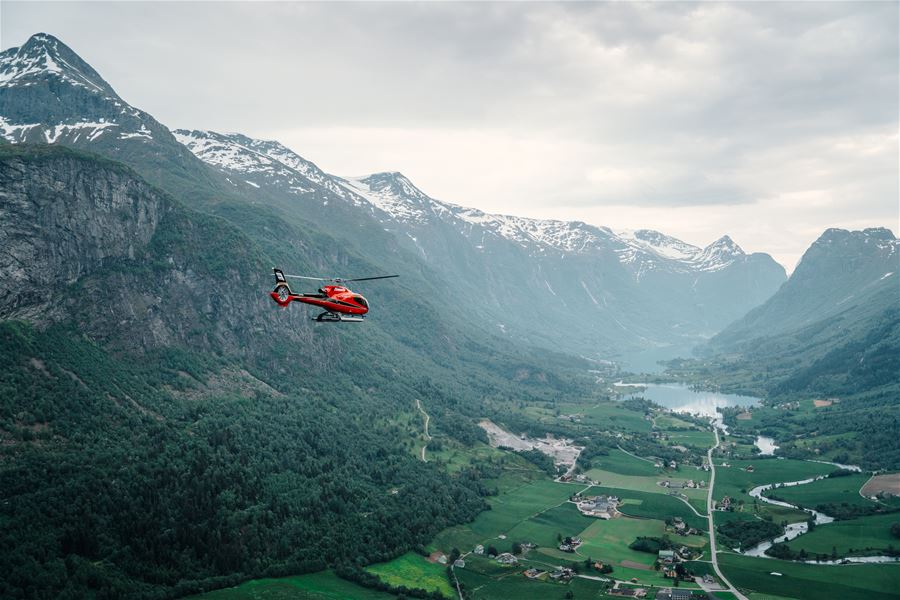 Fjord Helikopter - A red helicopter flies above a green valley with farmland, a lake, and snow-capped mountains under cloudy skies..