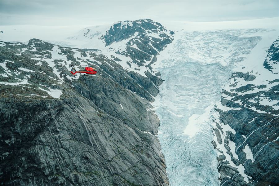 Fjord Helikopter - A red helicopter flies above a rocky, snow-dusted mountain ridge beside a wide glacier under an overcast sky..