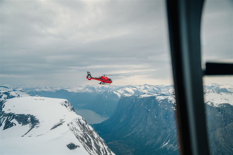 Fjord Helikopter flies above snow-covered mountains and a distant valley lake under an overcast sky, seen from inside another aircraft..