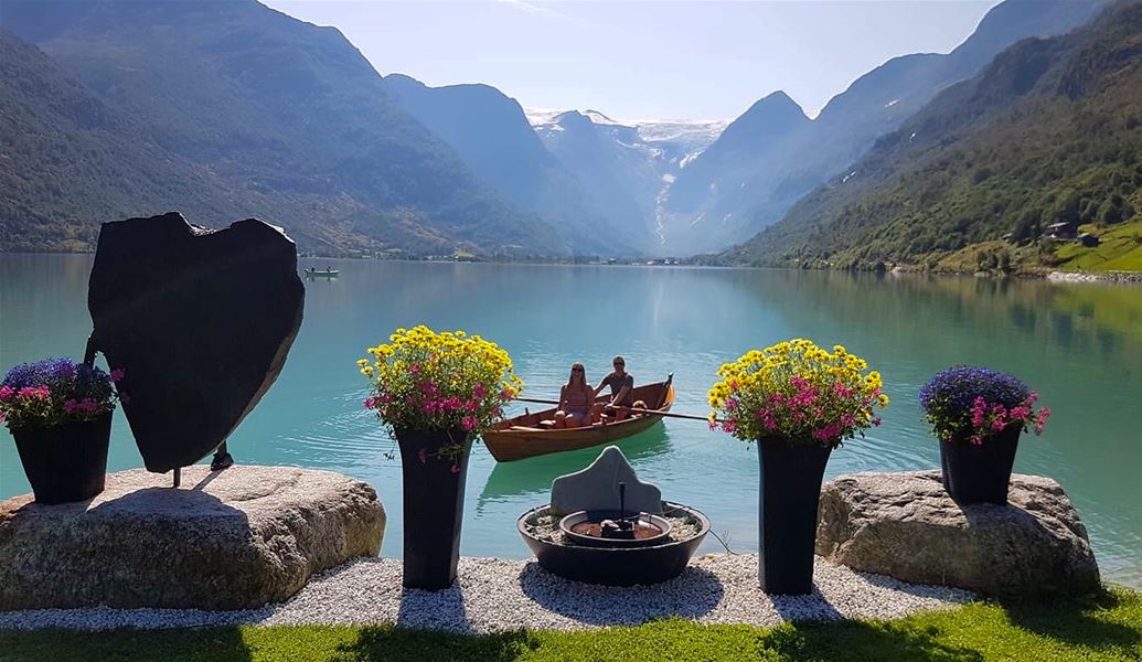 Couple in a wooden rowboat on a turquoise Oldenvatnetlake, framed by potted flowers and mountains with distant snow at Yrineset in Oldedalen..