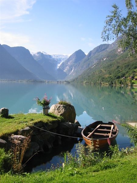 A wooden rowboat rests by a grassy shore on a calm Oldenvatnet lake, with forested mountains and distant snowfields under clear sky at Yrinset in Oldedalen, Norway..