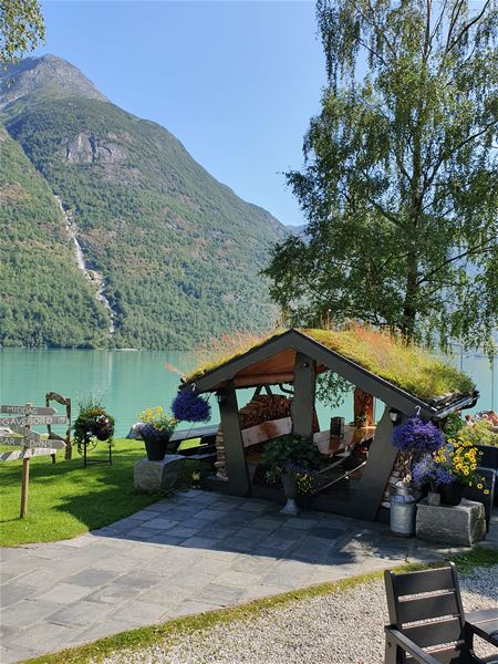 Small roofed shelter with potted flowers by a turquoise lake, with forested mountains and a waterfall in the distance..