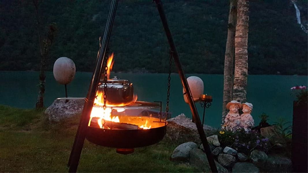 Metal pot and grill hang over a fire on a tripod in a grassy lakeside Oldenvatnet area at dusk, with trees behind at Yrinset in Oldedalen, Norway..