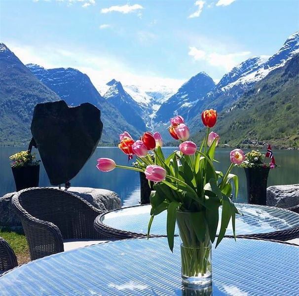 Vase of pink and red tulips on a glass patio table with wicker chairs, Oldenvatnet lake, and snowy mountains beyond at Yrinset in Oldedalen, Norway..