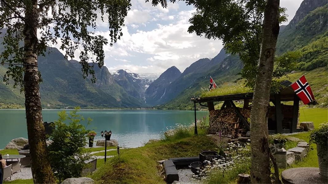 Lakeside scene framed by trees, with green mountains and a distant glacier, plus a small shed with flags at Yrinset in Oldedalen, Norway..