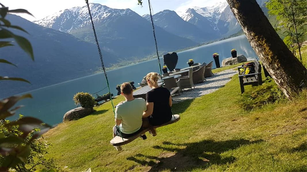 Two people sit on a wooden swing facing a blue Oldenvatnet lake, patio seating, and snowcapped mountains in sunlight at Yrinset in Oldedalen, Norway..